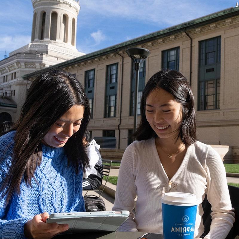 two students laughing at a table outside under a blue sky